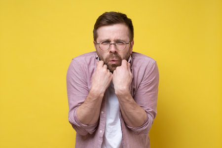 Young Caucasian man is very cold, he tries to wrap himself in a shirt, narrowed eyes and looks at the camera. Yellow background.の写真素材
