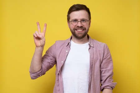 Joyful man in glasses and casual clothes shows the symbol of victory and looks at the camera smiling. Digit with two fingers.の写真素材