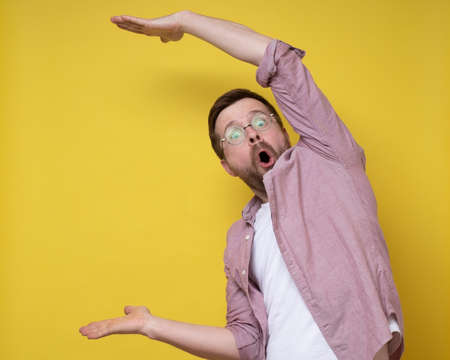 Surprised man in glasses shows a large size with hands, he exaggerates and looks at the camera in amazement. Copy space.の写真素材