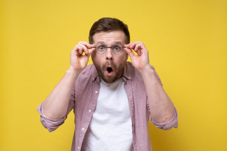 Shocked Caucasian man holds glasses, looks with amazement and open mouth. Yellow background.の写真素材