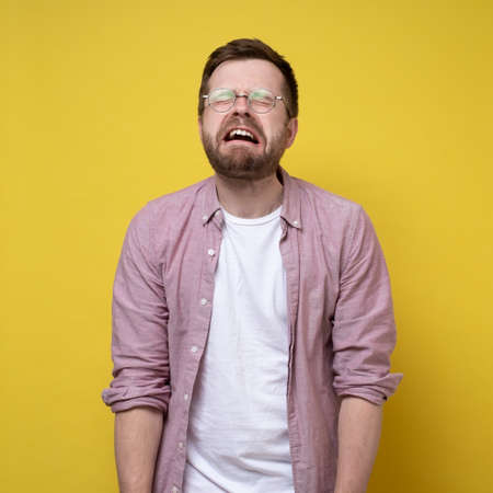 Sobbing, unhappy man in glasses and casual clothes, stands with hands down and eyes closed. Stress concept. Yellow background.の写真素材