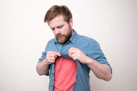 Focused bearded man unbuttons shirt after trying on. Shopping. White background.の写真素材