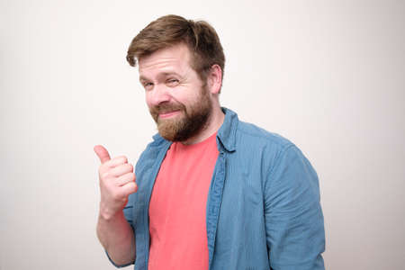 Dishonest Caucasian man points his thumb to the side and looks slyly. White background.の写真素材