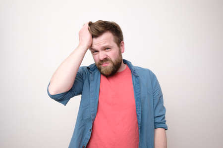 Young bearded man suffers from severe headache or is under stress, he holds forehead with hand and looks at the camera.の写真素材