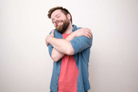 Charming bearded man hugs himself, he closed eyes and dreams of love, smiling sweetly. White background.の写真素材
