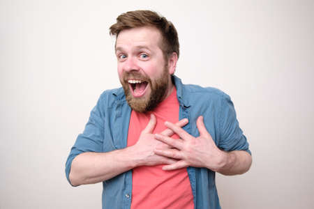 Joyful Caucasian man is surprised at a pleasant surprise, he smiles broadly, holds his hands over heart and looks at the camera.の写真素材
