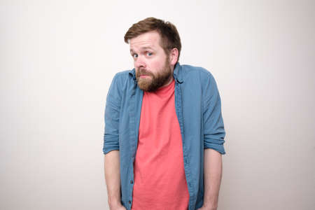 Bewilderment bearded man in casual clothes looks thoughtfully and in surprise, thrusting hands into pockets and lifting shoulders. White background.の写真素材