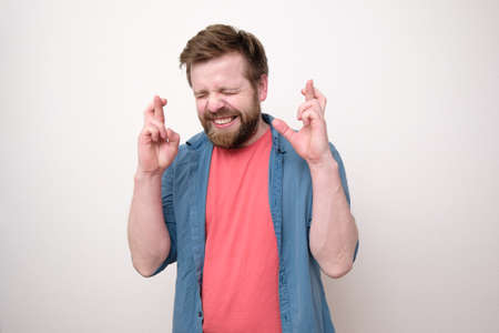 Cute bearded man really wants his wish to come true, he crossed fingers, gritted teeth emotionally and closed eyes.の写真素材