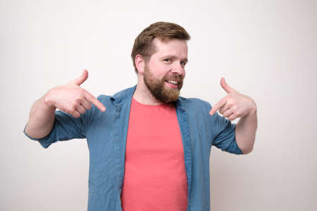 Funny bearded man in casual clothes persistently points at himself with index fingers, smiles sweetly and looks at the camera.の写真素材