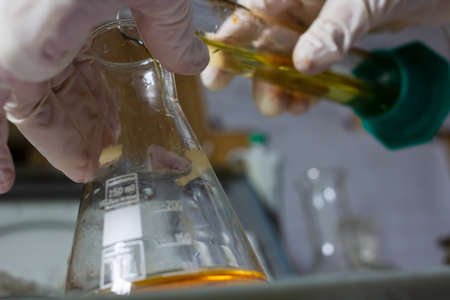 Gloved hands pour liquid from one flask into another, against a blurred background. Scientist is conducting an experiment in the laboratory.の写真素材
