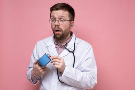 Sleepy doctor with glasses holds a mug of coffee and listens to it with a stethoscope. Pink background.の写真素材