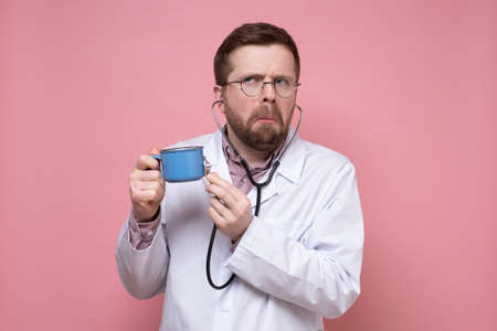 Perplexed, strange doctor with glasses holds a mug of coffee in hands and listens to it with a stethoscope. Pink background.の写真素材