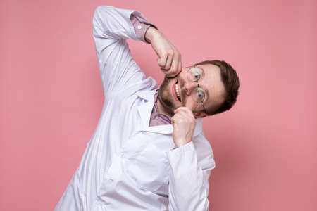 Funny doctor makes a smile by holding mouth with hands, he does not lose heart and entertains patients. Pink background.の写真素材