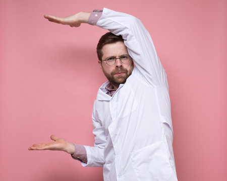 Serious doctor in round glasses shows something in a strange pose and looks sternly at the camera. Copy space. Pink background.の写真素材