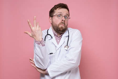 Young Caucasian doctor makes a hand gesture in bewilderment and looks in surprise. Pink background.の写真素材