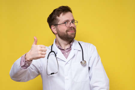 Cute, contented doctor shows an approving gesture, thumbs up and smiles friendly while looking into the camera.の写真素材