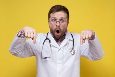Agitated doctor in a white coat with a stethoscope around neck points down with index fingers, looks indignantly with his mouth open.の写真素材