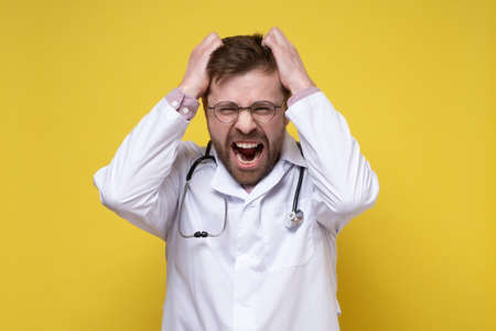Young doctor, stressed or furious, pulls his hair out and screams. Psychological concept. Yellow background.の写真素材
