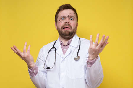 Puzzled doctor with a stupid expression on face and glasses on forehead, makes a confused gesture with hands. Yellow background.の写真素材