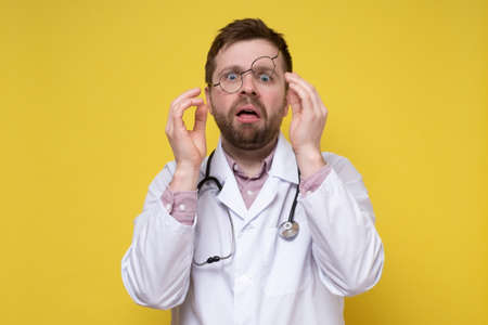 Terrified doctor in stress, he nervously adjusts glasses and looks in disbelief with a stupid expression on face. Yellow background.の写真素材