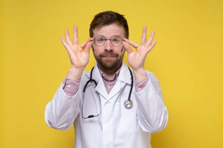 Cute doctor holds glasses in hands and looks through them, with a funny, benevolent expression on face. Yellow background.の写真素材