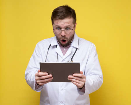 Frightened, shocked doctor looks at a tablet with an open mouth and big eyes. Yellow background.の写真素材