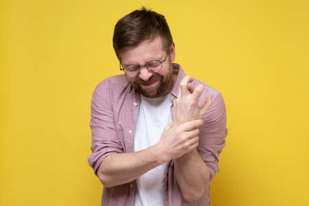Bearded man with glasses experiencing severe pain in hand, he held the wrist and closed eyes with a grimace of pain. Yellow background.の写真素材