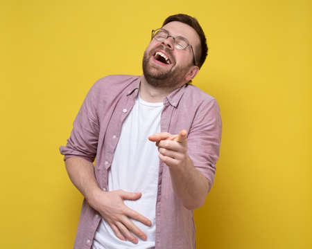 Man in round glasses and casual clothes laughs loudly and derisively, holds stomach and points finger at the camera.の写真素材