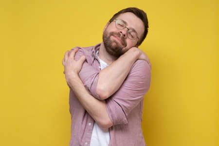 Cute, funny bearded man in round glasses hugs himself, he dreams of love or suffers from narcissism, closes eyes and smiles sweetly.の写真素材
