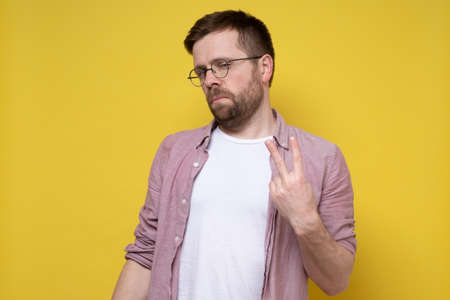 Serious man shows the number two on fingers or a victory sign and looks suspiciously at the camera. Yellow background.の写真素材