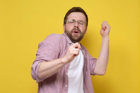 Caucasian man in a good mood, he dances making movements with hands and sings along with eyes closed. Yellow background.の写真素材