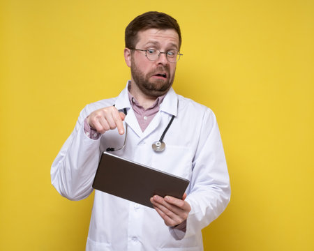 Surprised doctor holds a tablet in hands and excitedly points finger at the screen. Yellow background.の写真素材