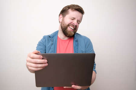 Cute man holds a laptop in hands and communicates via video communication, he laughs with eyes closed. Remote work or education.の写真素材