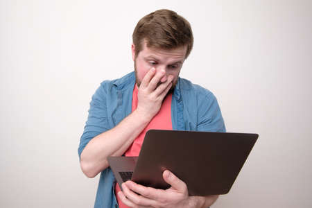 Upset man holds a laptop in hands and looks at the screen in bewilderment, covering part of face with hand. Remote work or education.の写真素材