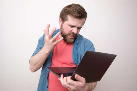 Angry man holds a laptop in hands, looks at the screen and swears with someone over video communication, making a hand gesture.の写真素材