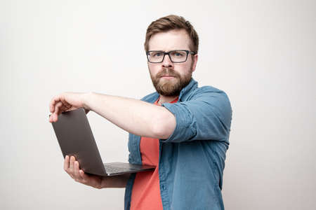 Serious, concentrated bearded man with glasses holds a laptop in hands and looks sternly at the camera. White background.の写真素材