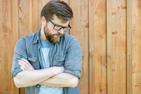 Pensive, attractive bearded man with glasses crossed his arms and looks down, against a background of a wooden wall. Copy space.の写真素材