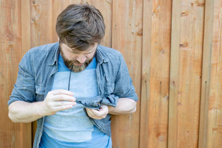 Concentrated man carefully cleans glasses from dust, wiping them with a shirt, against a background of a wooden wall. Copy space.の写真素材