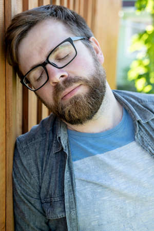 Caucasian, bearded man wearing glasses is tired and resting, leaning against a wooden wall and closing his eyes. Close-up.の写真素材