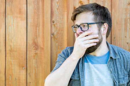 Man with glasses is tired, wants to sleep and yawns, covering mouth with hand, leaning against a wooden wall. Copy space. Close-up.の写真素材