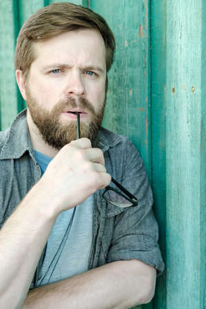 Caucasian, bearded man is thinking about something, holding glasses in mouth and looking thoughtfully.の写真素材