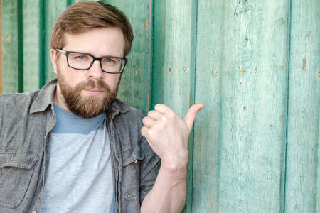 Attractive bearded man wearing glasses makes an approving gesture, thumb up or points to an empty wooden wall. Copy space.の写真素材