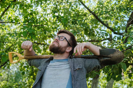 Bearded man in glasses holds a shovel on shoulders and looks up, dreamily, thoughtfully, against the backdrop of a garden.の写真素材