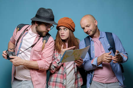 Tourists, two men and one woman, perplexedly look at the map and plan a route for a walk and sightseeing. Blue background.の写真素材
