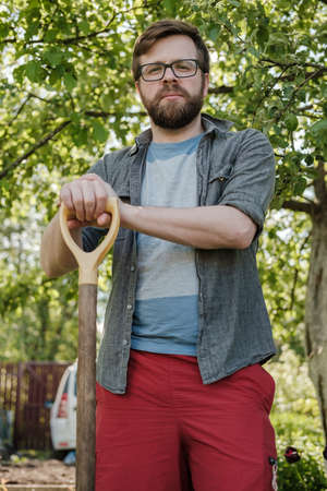 Serious man is leaning on the handle of a shovel and looking at the camera, in the garden, against the backdrop of a car and a fence.の写真素材