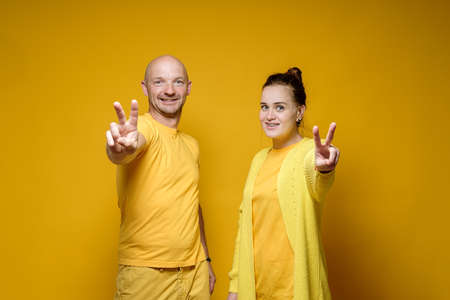 Smiling man and woman show victory symbol with fingers and look into the camera. Number two. Yellow background.の写真素材