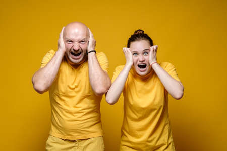 Man and woman are in stress, they cover their ears with their hands and scream hysterically. Yellow background.の写真素材