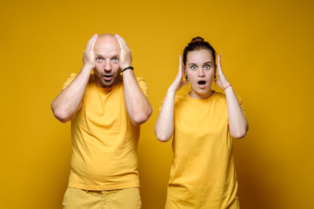 Man and the woman are shocked, they hold their heads with their hands and look fearfully with large eyes. Yellow background.の写真素材
