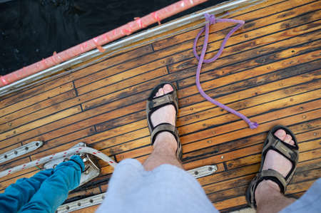 Male feet in sandals stand on the wooden deck of a yacht against the backdrop of dark water. Top view.の写真素材