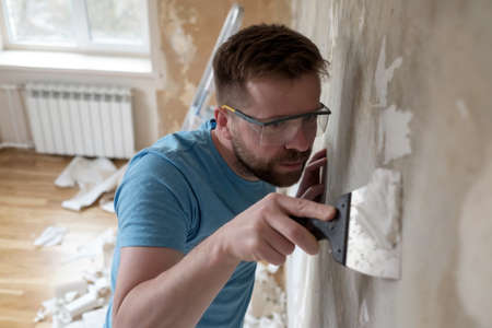 Concentrated man removes old wallpaper from the wall with a spatula in the room. Home repairs.の写真素材